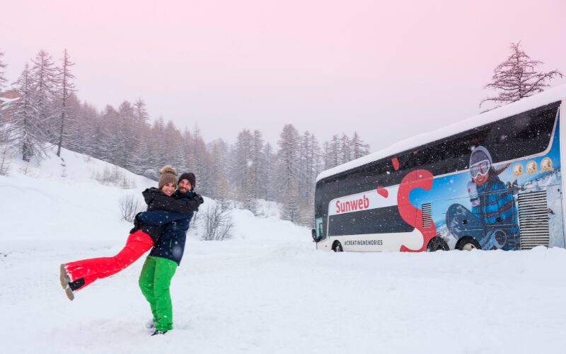 Couple hugging in the snow in front of a Sunweb coach after travelling to their ski holiday