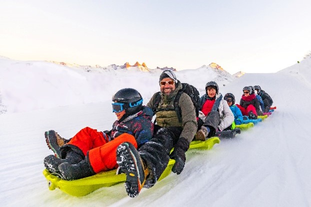 A group of smiling people ride snake gliss down a snowy slope, seated one after another on colourful linked sleds.