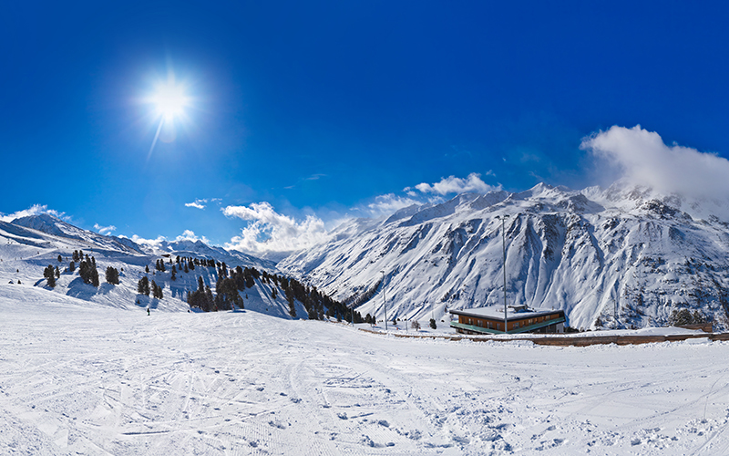 A snowy piste with a mountain backdrop under a clear blue sky, the bright sun shining directly into the camera.