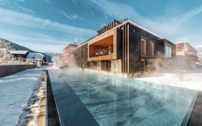 Outdoor heated pool with steam rising, set in a snowy landscape beside the wooden Falkensteiner Hotel Kronplatz in Italy.