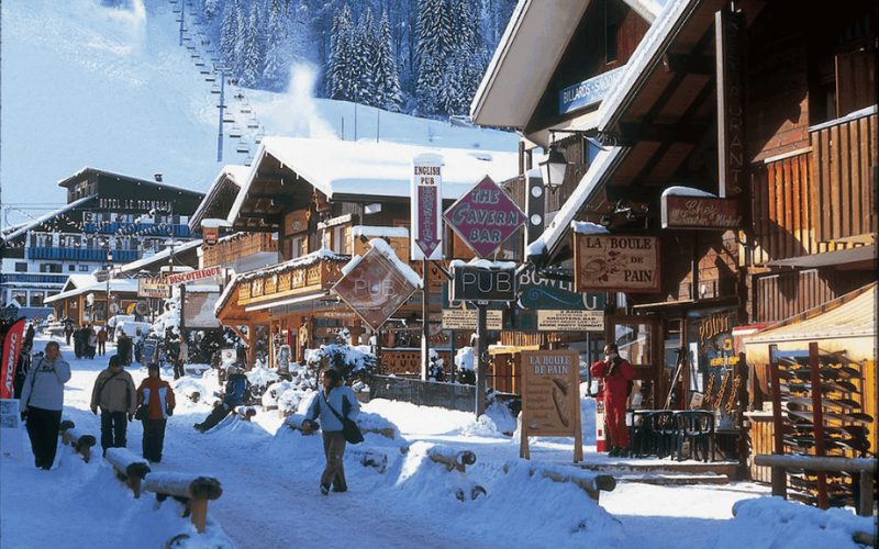 Alpine village street in Morzine, France, with wooden chalets and pedestrians, and a chairlift rising toward the mountain in the background.