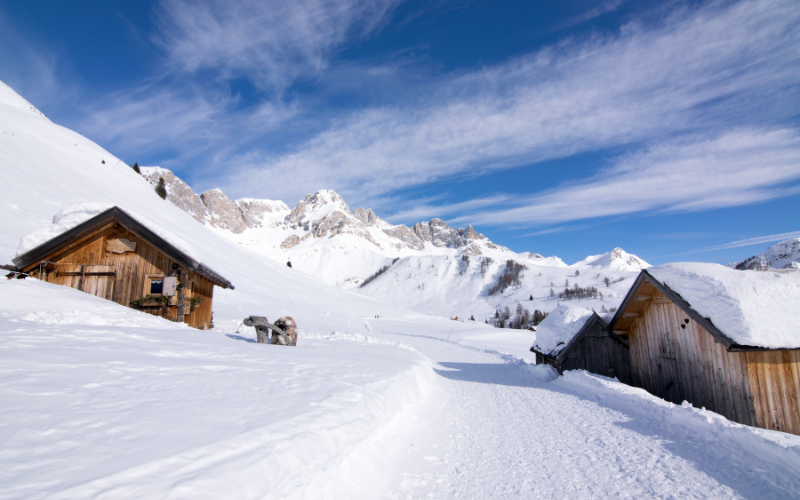 A piste on the Sella Ronda route with snow‑covered houses beside it and snow‑covered mountains in the background.